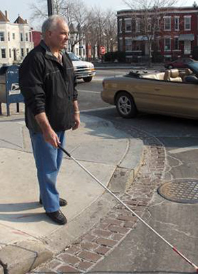 photo shows the man with his cane in front, starting to walk into the crosswalk to cross the street.  The cars in the street beside him are moving forward.